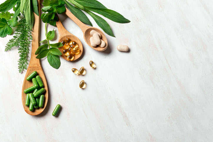 Supplements on wooden spoons next to green vegetables and plants